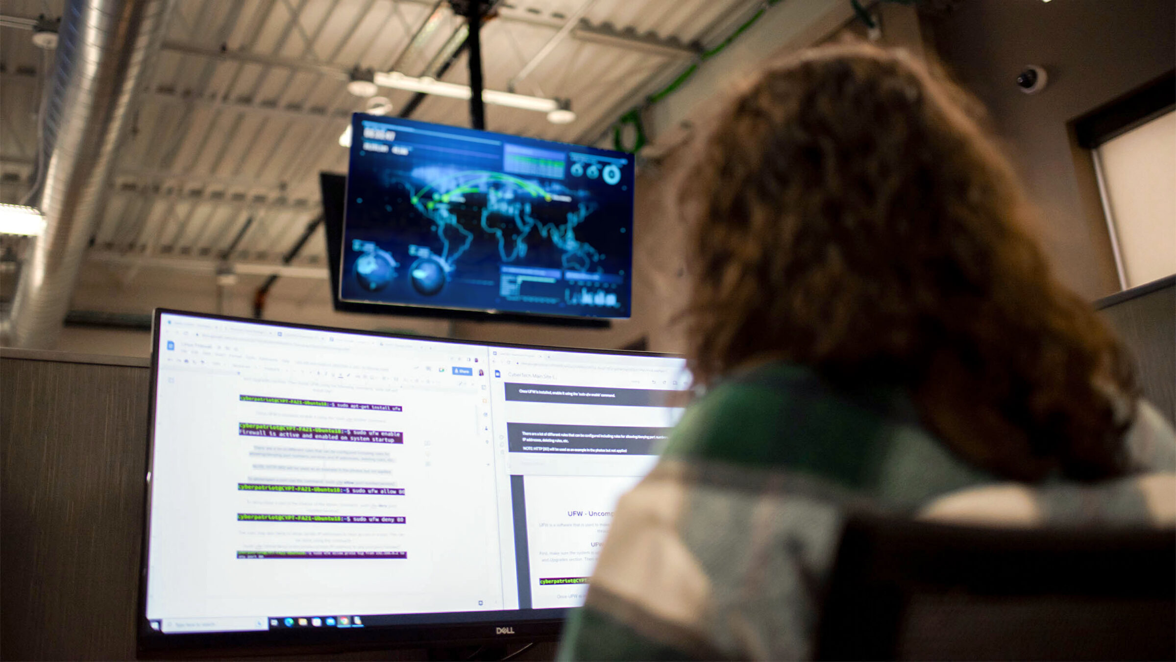 computer & digital forensics student writing code on a computer; threat assessment software running on an overhead monitor