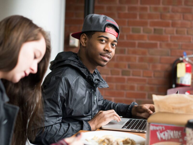 A student working on their laptop while eating in IDX looking at the camera