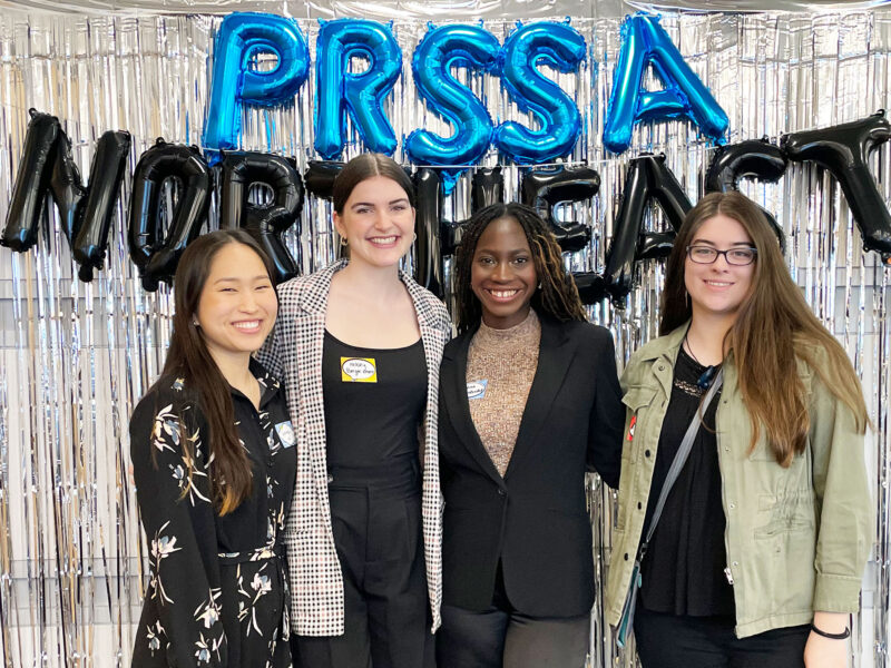 students posing in front of a silver background with PRSSA Northeast written in blue and black balloon letters