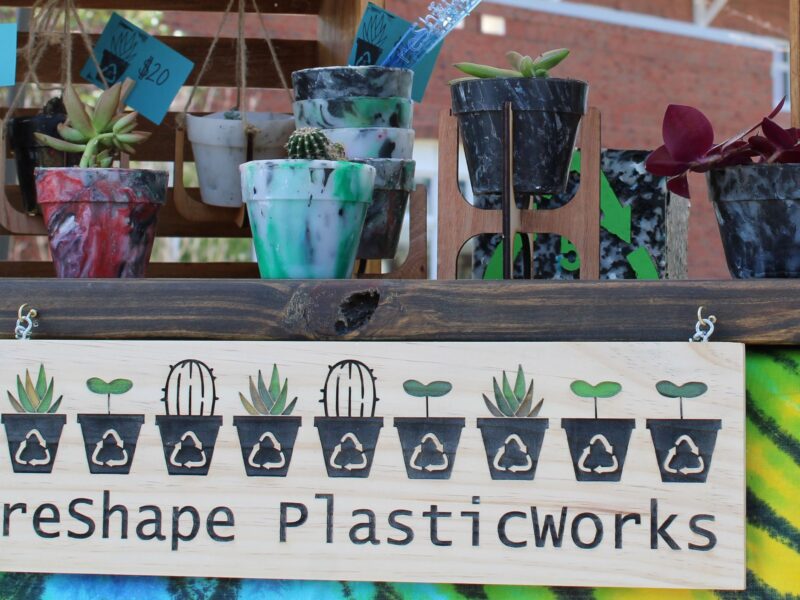A collection of plastic planters and plant pots on a wooden shelf above a company logo reading reShape PlasticWorks